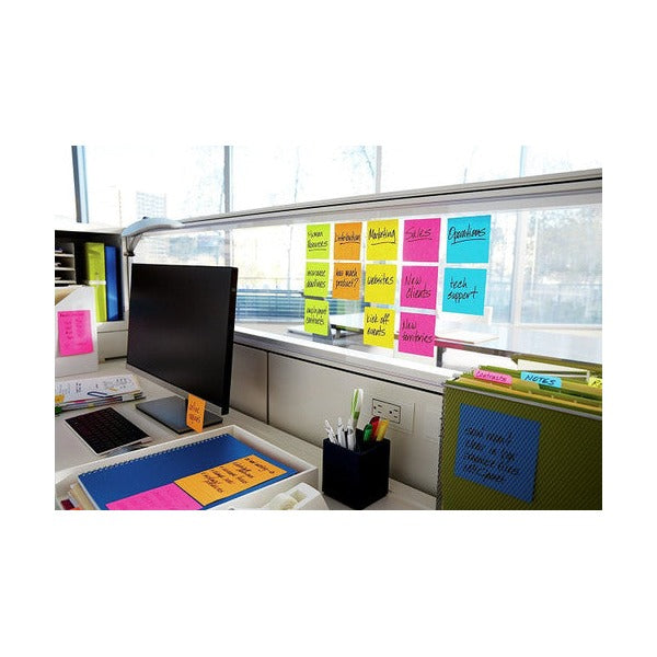 Office window covered in a grid of coloured sticky notes with handwritten words, beside a monitor and desk tidy