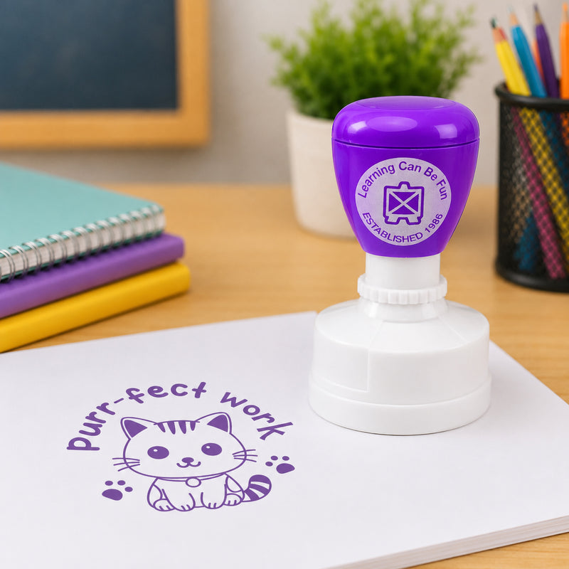 Purple merit stamp shown in a classroom setting with notebooks and educational supplies. The clear kitten themed impression helps teachers recognise quality work, positive learning behaviours and academic success.