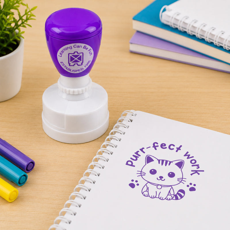 Teacher reward stamp positioned next to colourful markers and exercise books. The Purr-fect Work design features a friendly kitten illustration that provides fun and encouraging feedback for classroom activities and assignments.