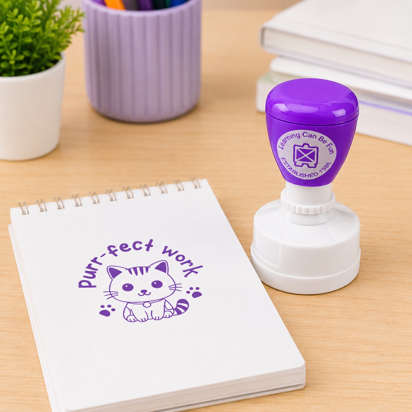 Learning Can Be Fun Purr-fect Work Merit Stamp displayed beside a spiral notebook and classroom stationery. The purple self inking stamp creates a cute kitten themed reward impression designed to celebrate excellent student work and achievement.