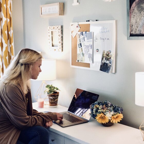 Quartet Basics combination board hung above a home desk while a woman works at a laptop below it, with papers pinned to the cork and handwritten notes on the white surface