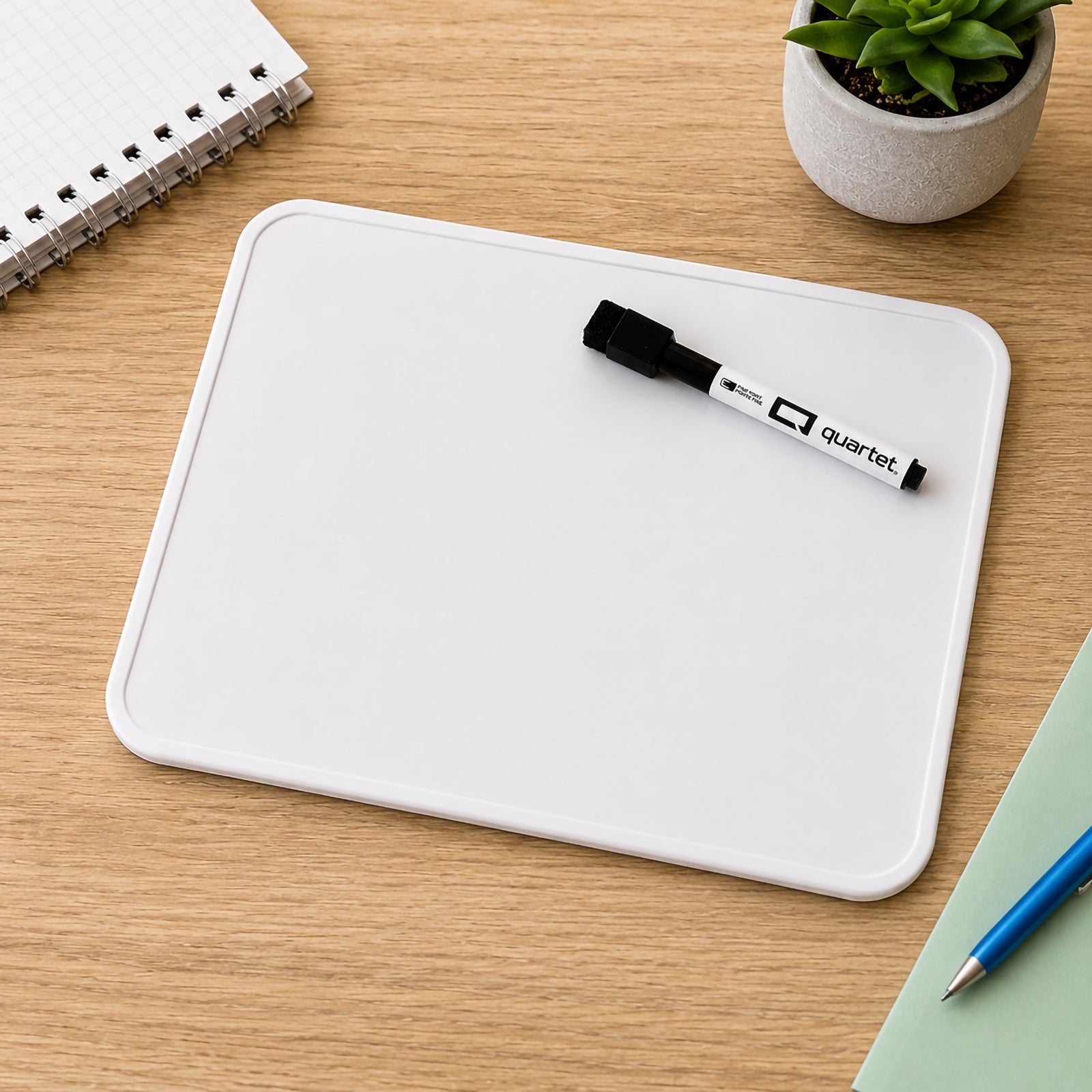 Top view of the Quartet Double Sided Magnetic Lap Board 210 x 300mm placed on a study desk beside a notebook, stationery and plant. The clean reusable writing surface and included dry erase marker demonstrate an organised learning setup for students and teachers.