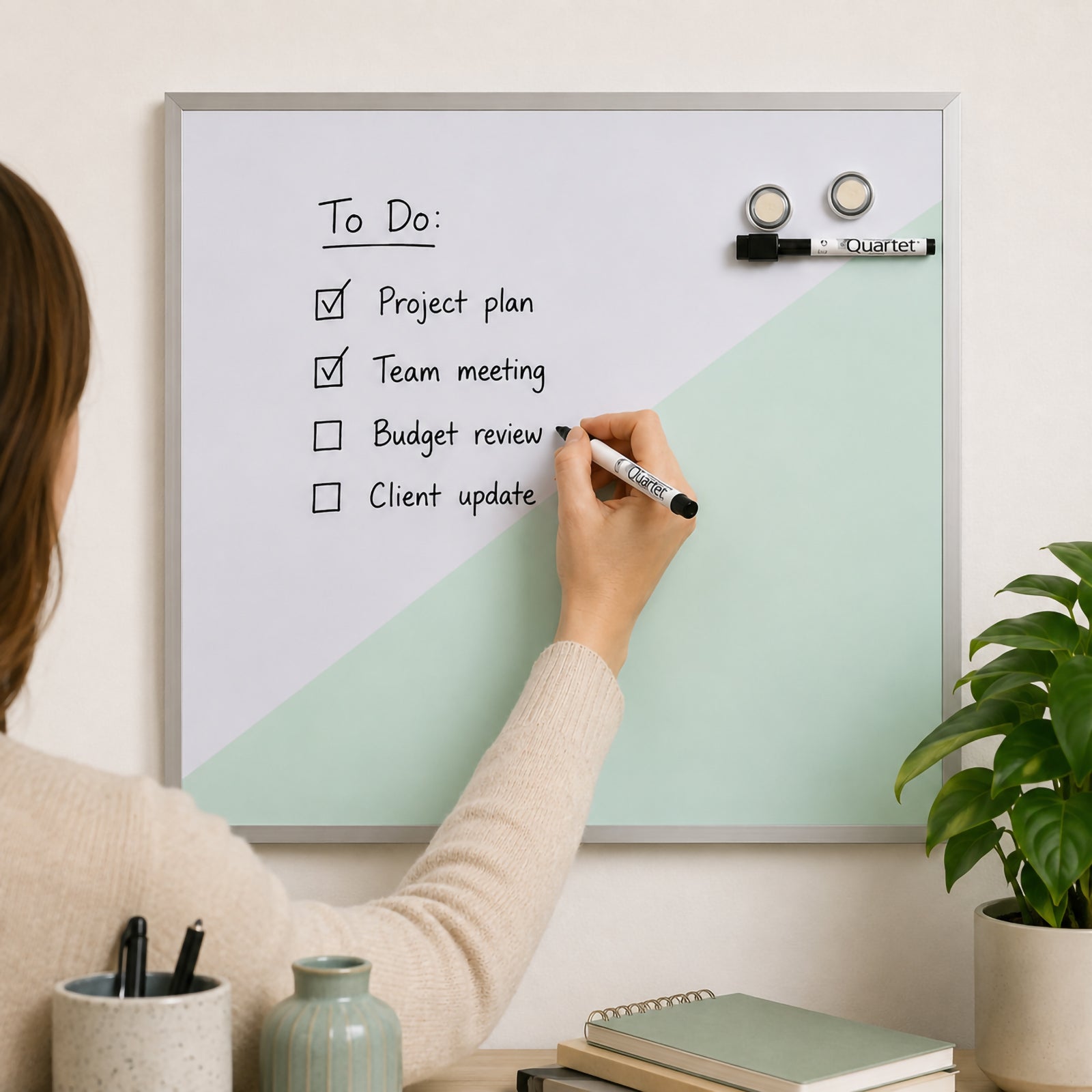 Lifestyle image showing the Quartet Geo Magnetic Whiteboard Teal 430 x 580mm mounted on a home office wall while a user updates a daily task list. The stylish teal geometric design complements modern décor, demonstrating practical organisation for schedules, reminders and project planning.