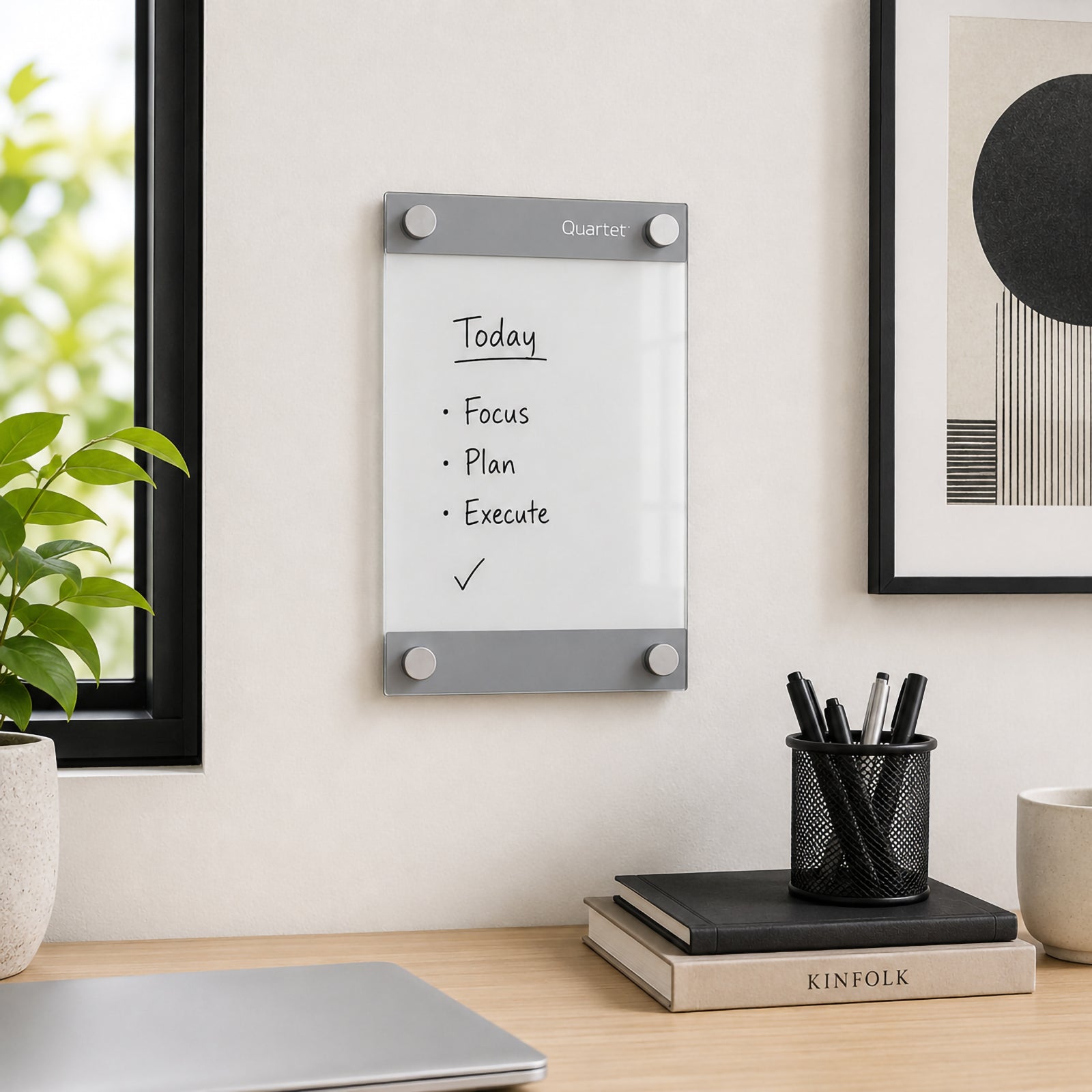 Quartet Infinity Glassboard Custom 215 x 279mm installed vertically in a bright contemporary office beside a window and framed artwork. The compact glassboard displays a simple focus list, while the clear writing surface and silver mounting hardware complement modern decor without creating unnecessary visual clutter.