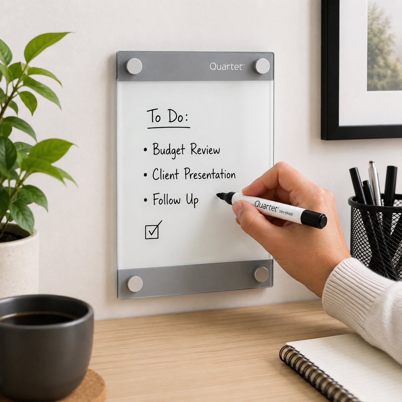 Quartet Infinity Glassboard Custom 215 x 279mm mounted above a timber desk while a person writes a compact task list with a Quartet dry erase marker. The clear magnetic glass surface, silver mounting panels and frameless floating design create a polished planning area suited to home offices, study spaces and professional workstations.