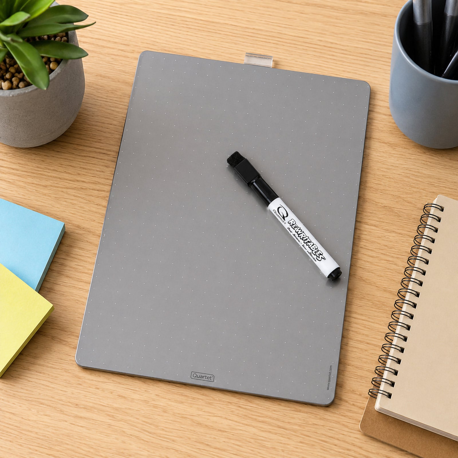 Top view of the Quartet Magnetic Whiteboard Tile Silver 216 x 280mm resting on a wooden desk beside notebooks, sticky notes and office stationery. The blank metallic dry erase surface highlights its clean reusable design for home, study and workplace organisation.