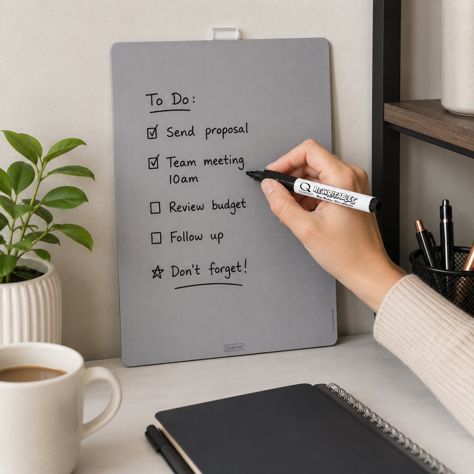 Lifestyle image showing the Quartet Magnetic Whiteboard Tile Silver 216 x 280mm positioned on a home office shelf with a handwritten task list. A hand writes on the smooth dry erase surface using the included marker, demonstrating everyday organisation for reminders, schedules and planning.