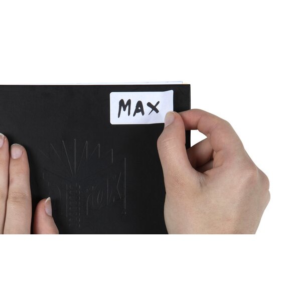 Hand pressing a white QuikStik label handwritten with the name MAX onto the cover of a black book