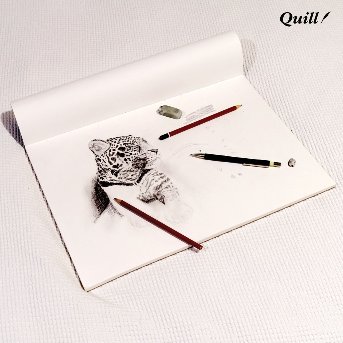 Quill Sketch Pad A3 open with the cover rolled back, a pencil drawing of a leopard on the white cartridge sheet and pencils and an eraser beside it