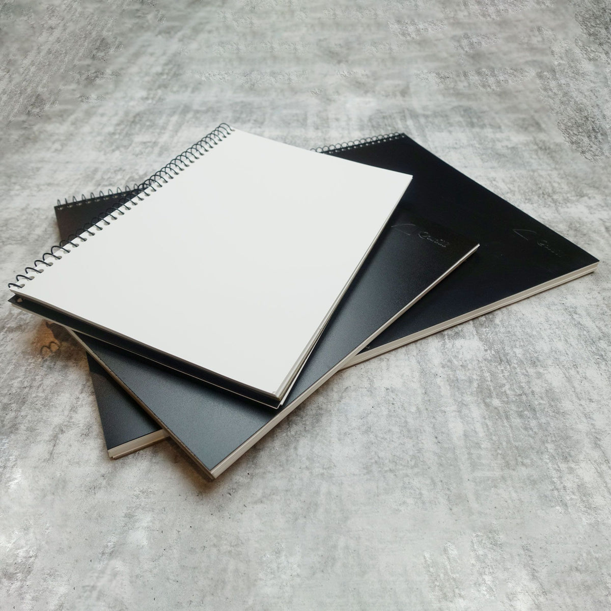Two Quill Visual Art Diaries A4 with black covers stacked on a grey surface, the top diary open to a blank white cartridge page