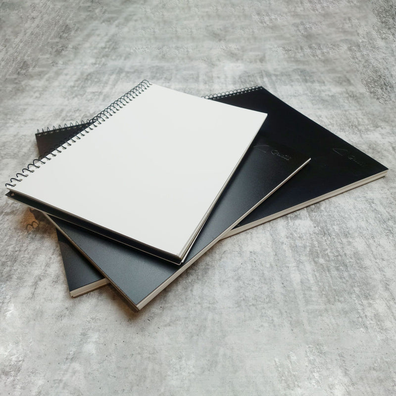 Two Quill Visual Art Diaries A4 with black covers stacked on a grey surface, the top diary open to a blank white cartridge page