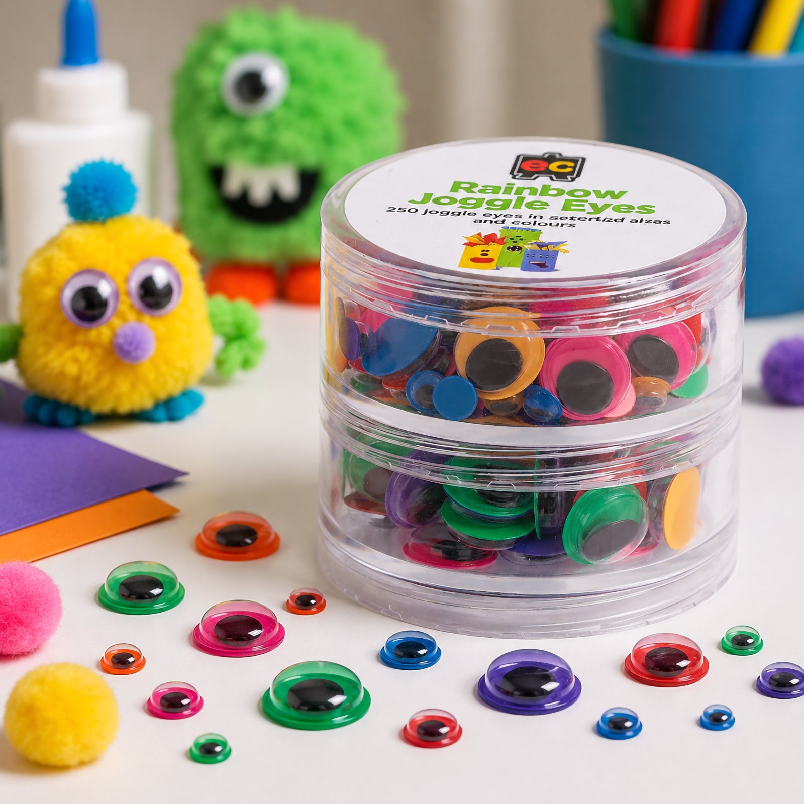 Close up of Educational Colours Rainbow Joggle Eyes stored in a clear reusable container. Various sizes and bright colours including pink, blue, green, yellow and purple are scattered across a craft table with pom pom characters and art supplies in the background.