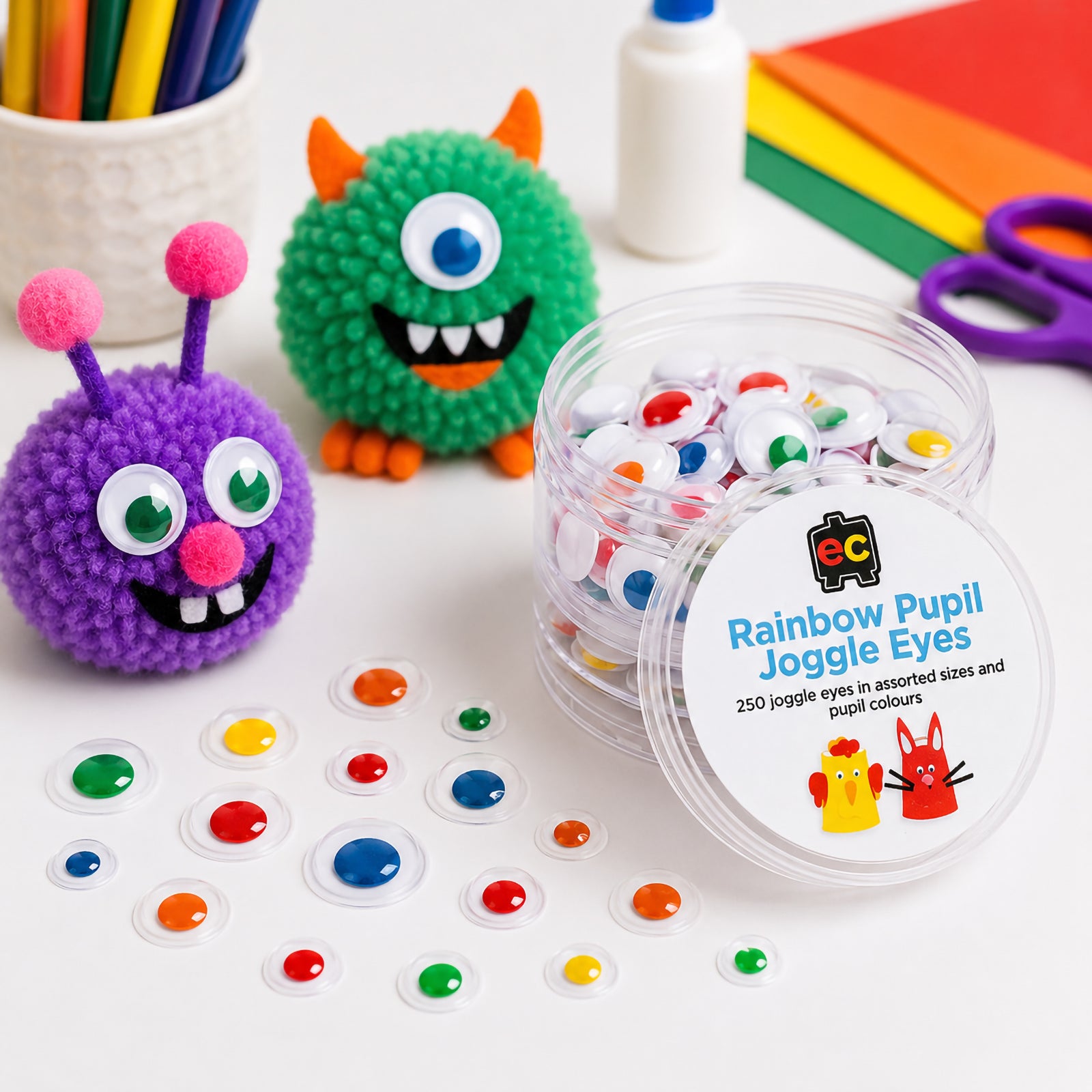 Educational Colours Rainbow Pupil Joggle Eyes displayed with colourful pom pom monster characters on a craft table. The image showcases assorted eye sizes with bright coloured pupils in red, blue, green, yellow and orange. A reusable storage container sits alongside scattered craft eyes, coloured paper, glue and scissors, demonstrating creative classroom and home craft applications for imaginative art projects and character creation.
