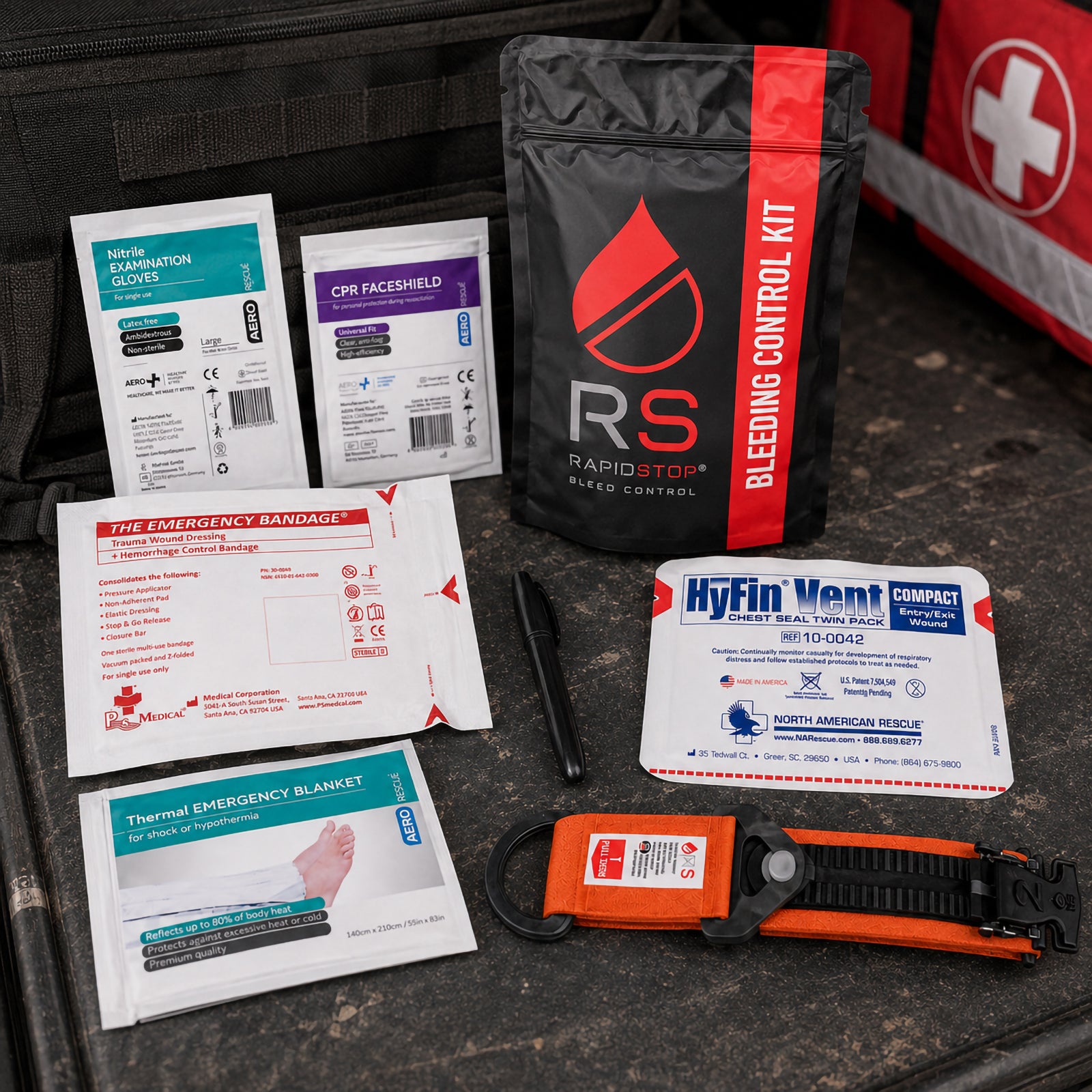 RapidStop Medium Bleeding Control Kit displayed beside emergency response bags in a workplace setting. The black and red pouch stands behind the tourniquet, trauma dressing, HyFin Vent chest seal, emergency blanket, nitrile gloves, CPR faceshield and marker, demonstrating the broader trauma equipment included in this portable configuration.