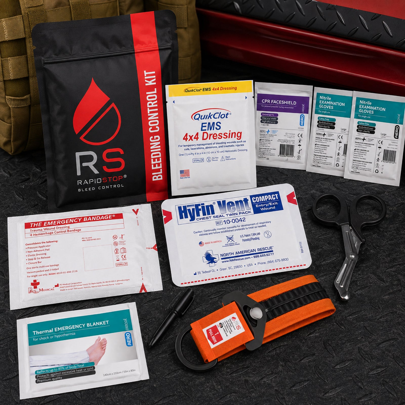 RapidStop Medium trauma response equipment presented in an industrial setting with the black and red bleeding control pouch positioned behind the QuikClot EMS 4x4 Dressing. The image also shows the HyFin Vent Compact chest seal, emergency bandage, thermal blanket, CPR faceshield, nitrile gloves, trauma shears, marker and RapidStop tourniquet.