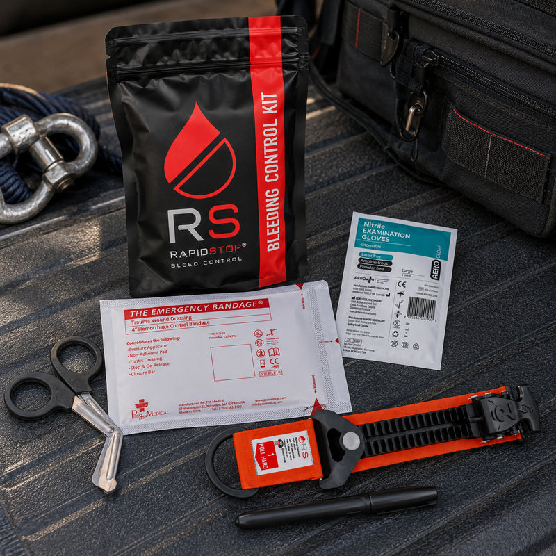 RapidStop Small bleeding control kit displayed on a vehicle cargo surface beside workplace response equipment. The compact black and red RapidStop pouch is positioned with the orange tourniquet, emergency trauma bandage, nitrile gloves, trauma shears and marker, demonstrating a practical configuration for mobile and workplace emergency preparedness.