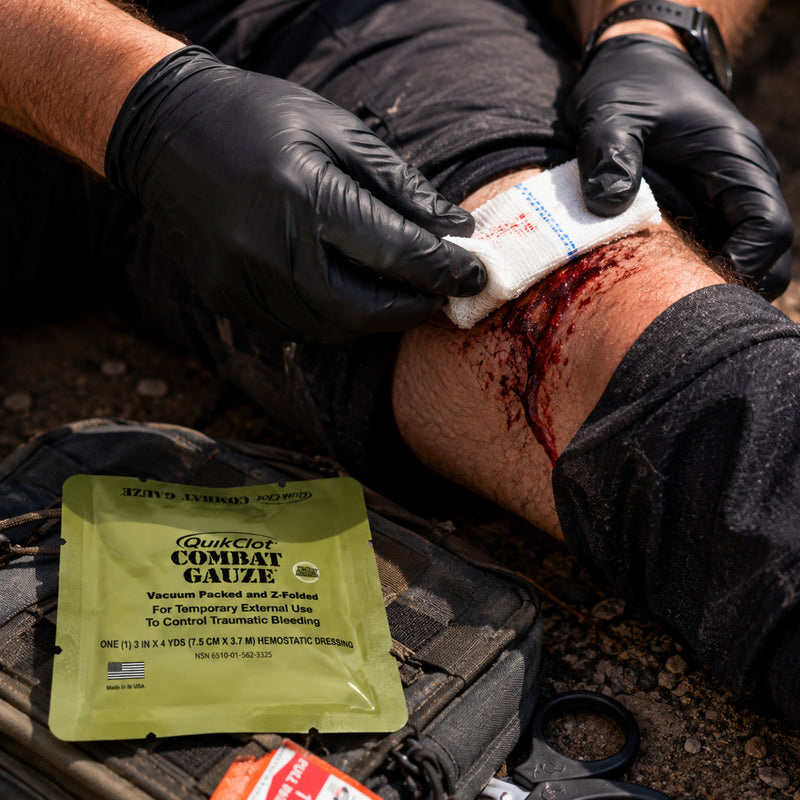 Close view of a simulated severe leg injury being treated with folded gauze by a responder wearing black examination gloves. A QuikClot Combat Gauze package is visible beside the casualty, illustrating the haemostatic gauze in an emergency trauma response setting with additional RapidStop bleeding control equipment nearby.