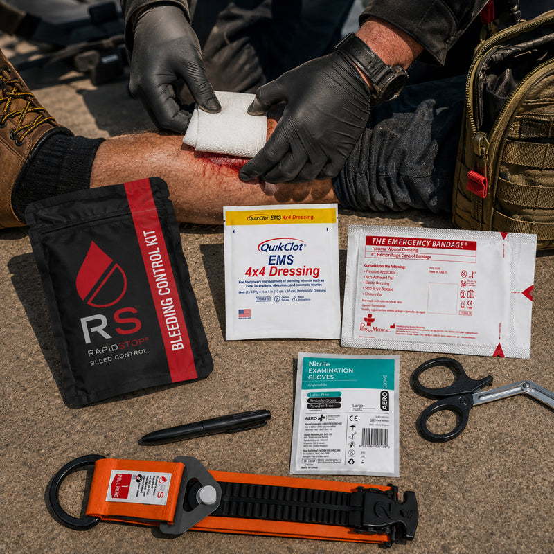 RapidStop Bleed Control Small Pack displayed during a simulated outdoor emergency response as a gloved responder applies a dressing to a leg injury. Visible kit contents include the RapidStop tourniquet, QuikClot EMS 4x4 Dressing, emergency trauma bandage, nitrile examination gloves, trauma shears, marker and black RapidStop pouch.