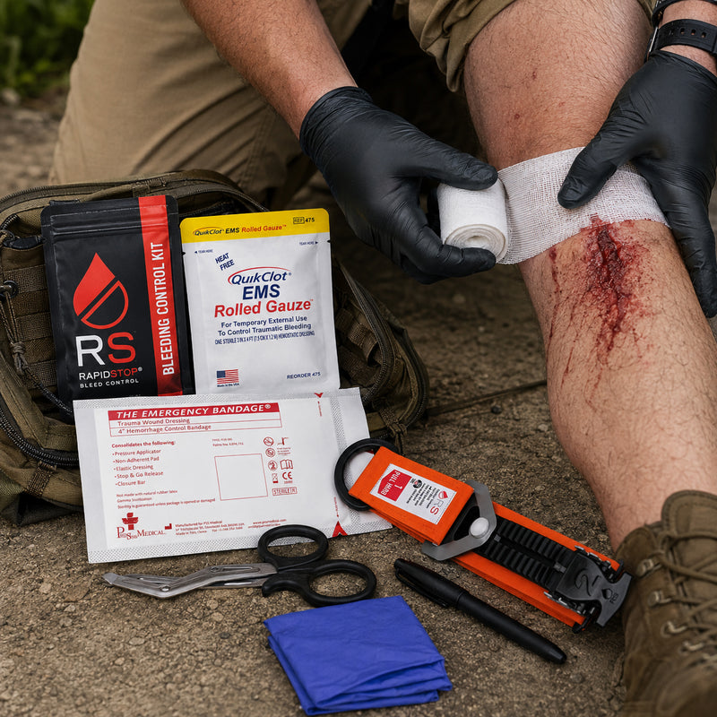 RapidStop Bleed Control Small Pack shown during a simulated outdoor trauma response, with a responder applying gauze to a leg wound. RapidStop tourniquet, QuikClot EMS Rolled Gauze, emergency trauma bandage, trauma shears, marker and examination gloves are arranged nearby for rapid access.