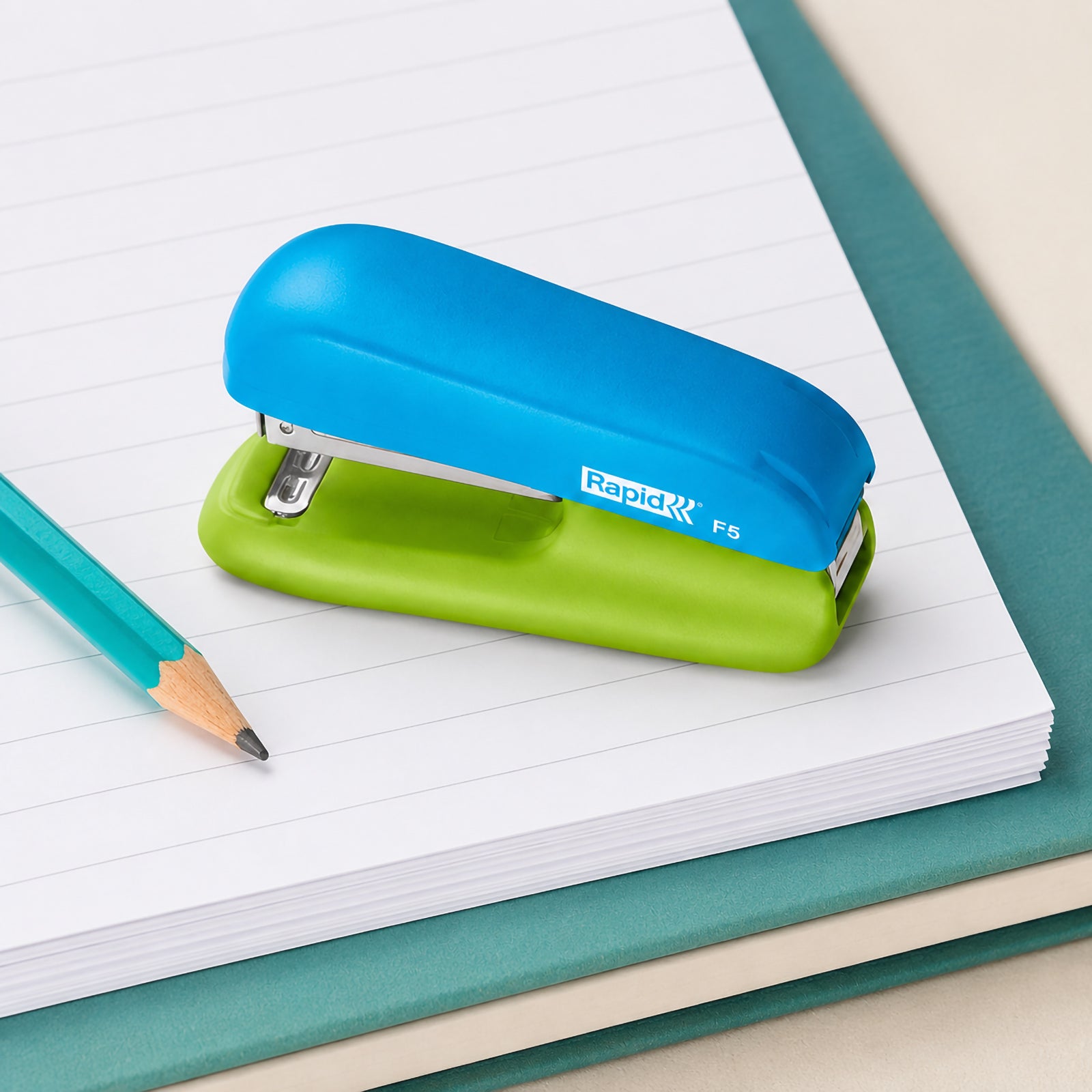 The Rapid F5 Mini Stapler rests on a lined notebook beside a sharpened pencil and writing pad. The close up composition highlights the compact blue and green design, making it ideal for study desks, classrooms and portable stationery collections.