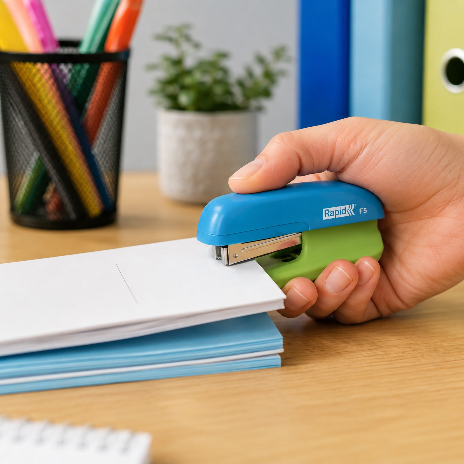 A lifestyle image showing a person using the Rapid F5 Mini Stapler in blue and green to staple a small stack of documents on a wooden desk. Colourful stationery, folders and office accessories in the background highlight its comfortable grip and suitability for everyday office, school and home use.