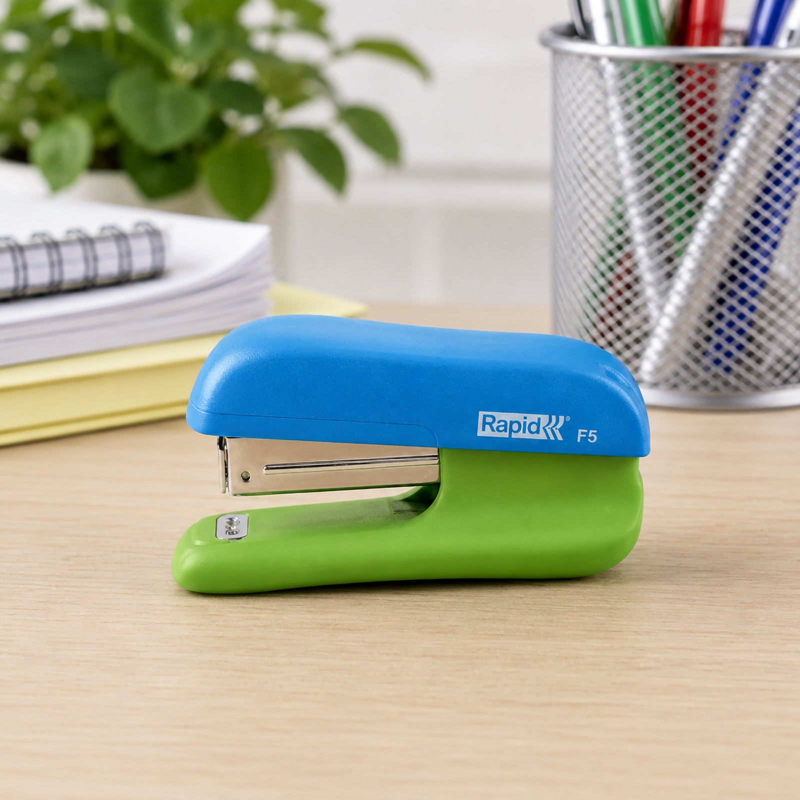 A desktop lifestyle scene featuring the Rapid F5 Mini Stapler positioned neatly on a tidy workspace with notebooks, a pen holder and office supplies. The image showcases the stapler's compact footprint and colourful finish in a modern office environment.