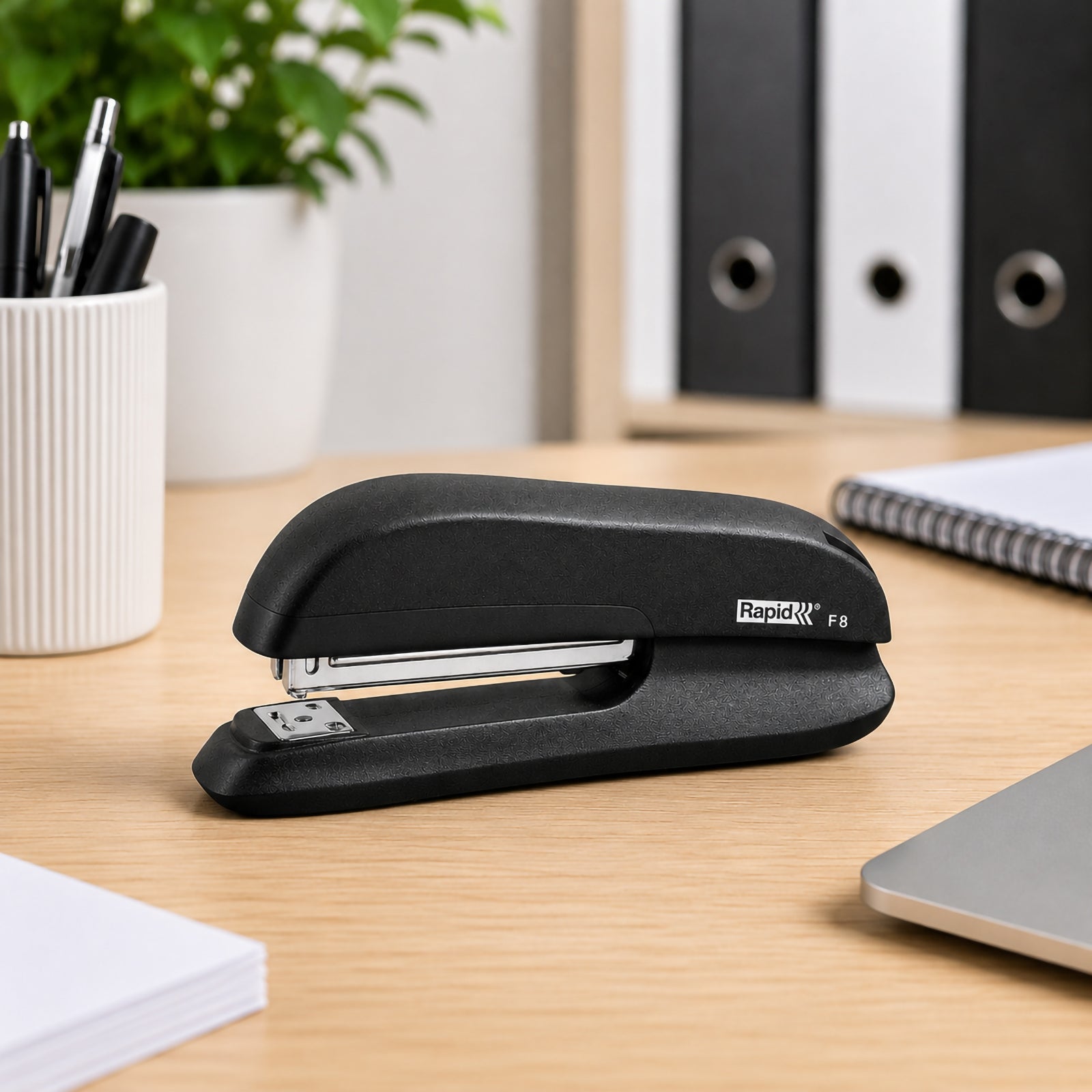 The Rapid F8 Full Strip Stapler is positioned on a tidy office desk surrounded by a laptop, stationery holder, notebooks and filing folders. The image showcases the stapler's professional black finish and compact full strip design in a contemporary workplace.