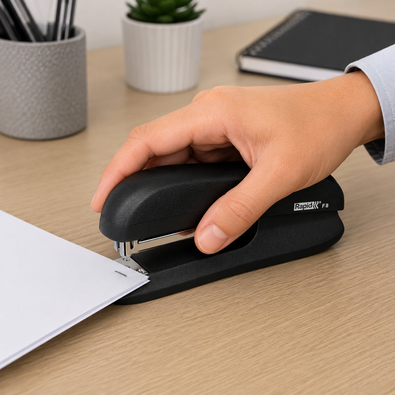 A close up lifestyle image showing a hand comfortably using the Rapid F8 Full Strip Stapler in black to staple a stack of office documents on a modern wooden desk. The workspace includes stationery, notebooks and office accessories, highlighting the ergonomic design and dependable everyday performance.