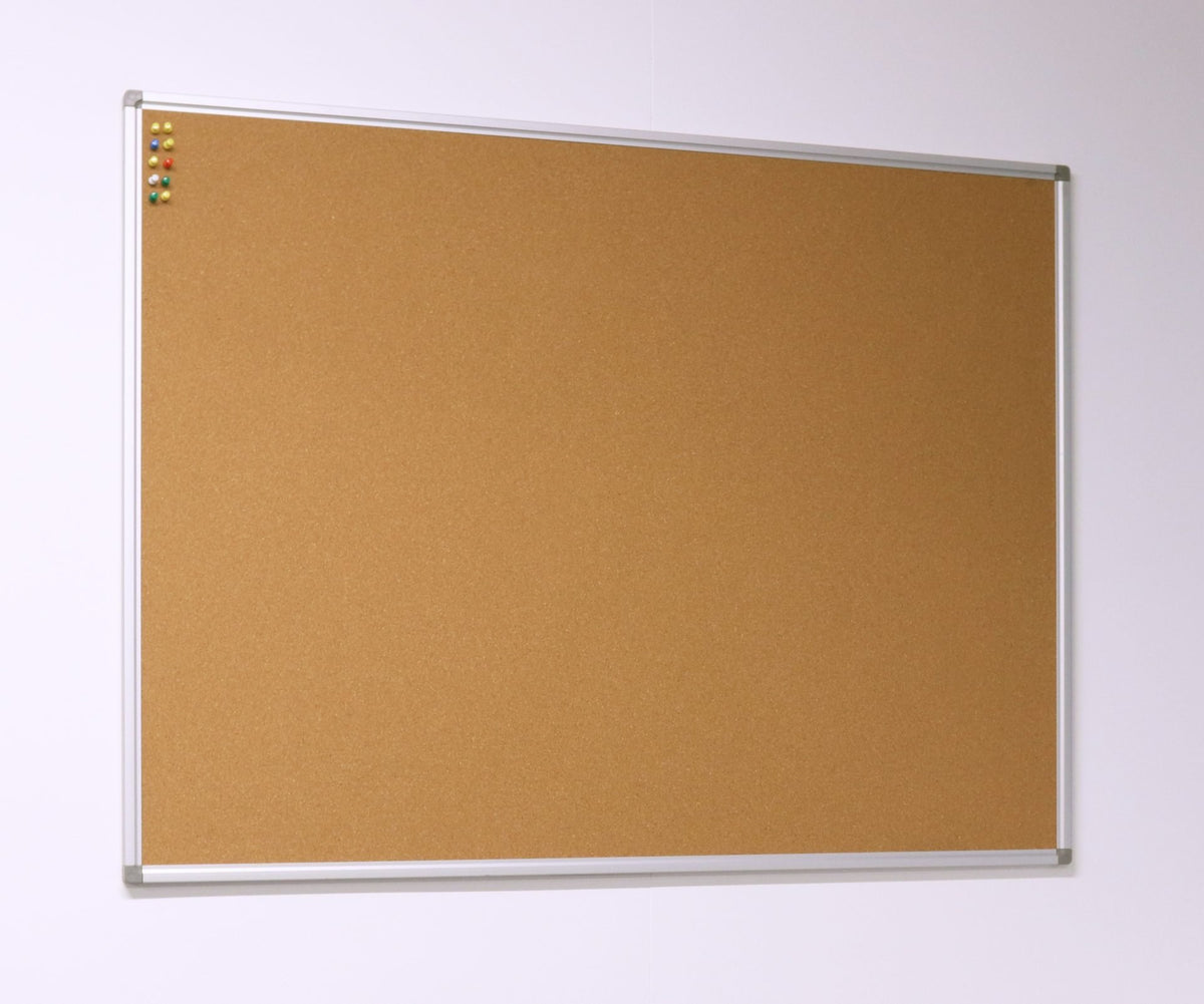 Rapidline Standard Corkboard hung on a pale wall and seen from an angle, with an aluminium frame and pins clustered in the top left corner.