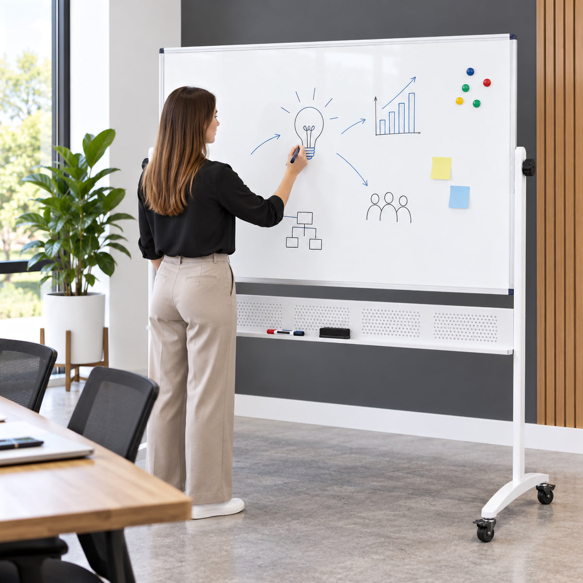 Rapidline Standard Mobile Magnetic Whiteboard 1500 x 1200mm being used during a workplace presentation in a modern meeting room. A person writes ideas, charts and planning diagrams across the large magnetic surface while the aluminium framed board stands securely on its mobile castor base, demonstrating its suitability for collaborative meetings and presentations.
