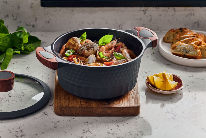 A Resto Capella casserole filled with clams and prawns, its glass lid set to one side, showing the 3D-textured exterior and wood-grain side handles (Resto lifestyle image)