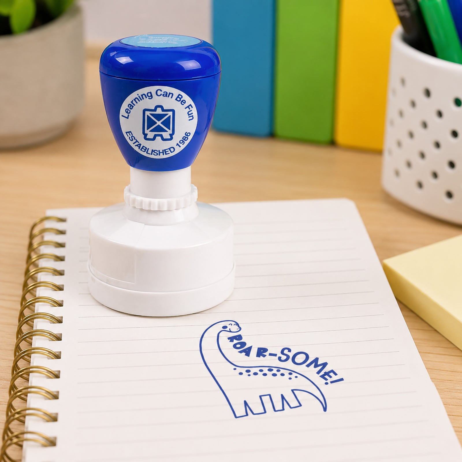 Blue Roar-some merit stamp positioned on a lined notebook with colourful classroom supplies in the background. The friendly dinosaur design encourages positive reinforcement and helps teachers celebrate academic success in an exciting way.