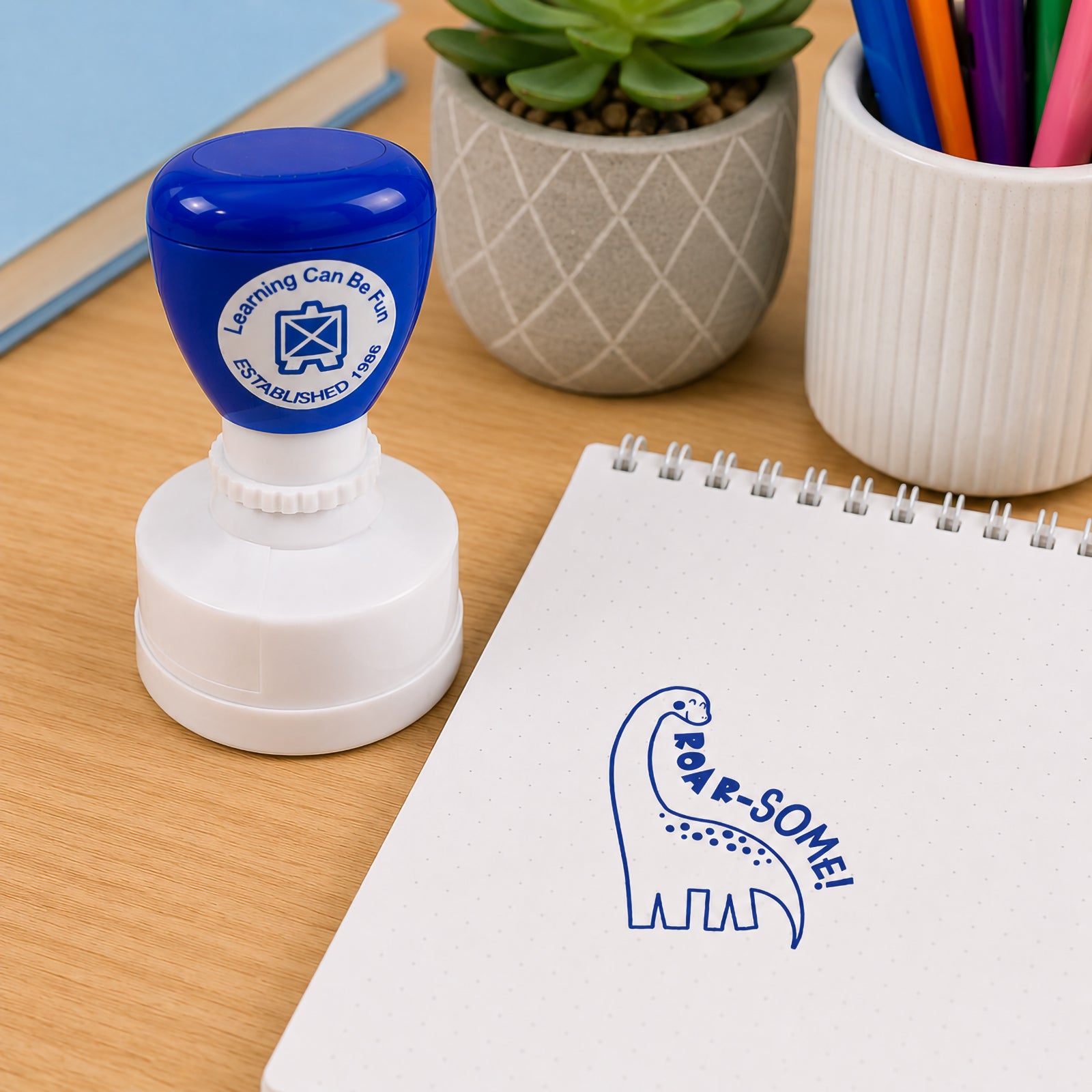 Learning Can Be Fun Roar-some Merit Stamp displayed beside a notebook on a classroom desk. The self inking teacher stamp creates a bold dinosaur themed impression in blue ink, making it a fun and engaging way to recognise student achievement and effort.