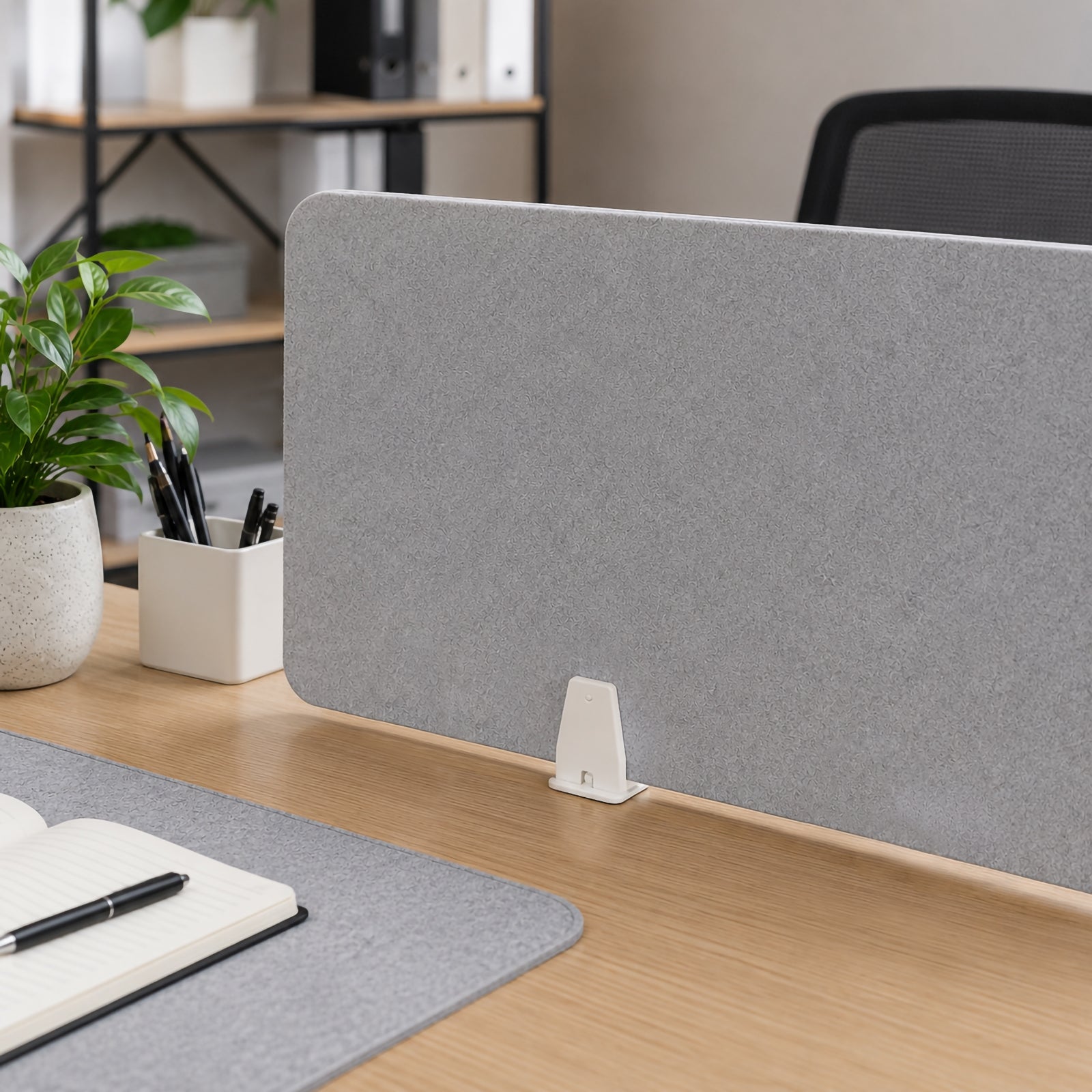 Close up lifestyle view of a SANA Type 1 matte white top mount clamp holding a grey PET desk screen on a timber desktop, with office plants, stationery and an open notebook creating a clean professional workspace setting.
