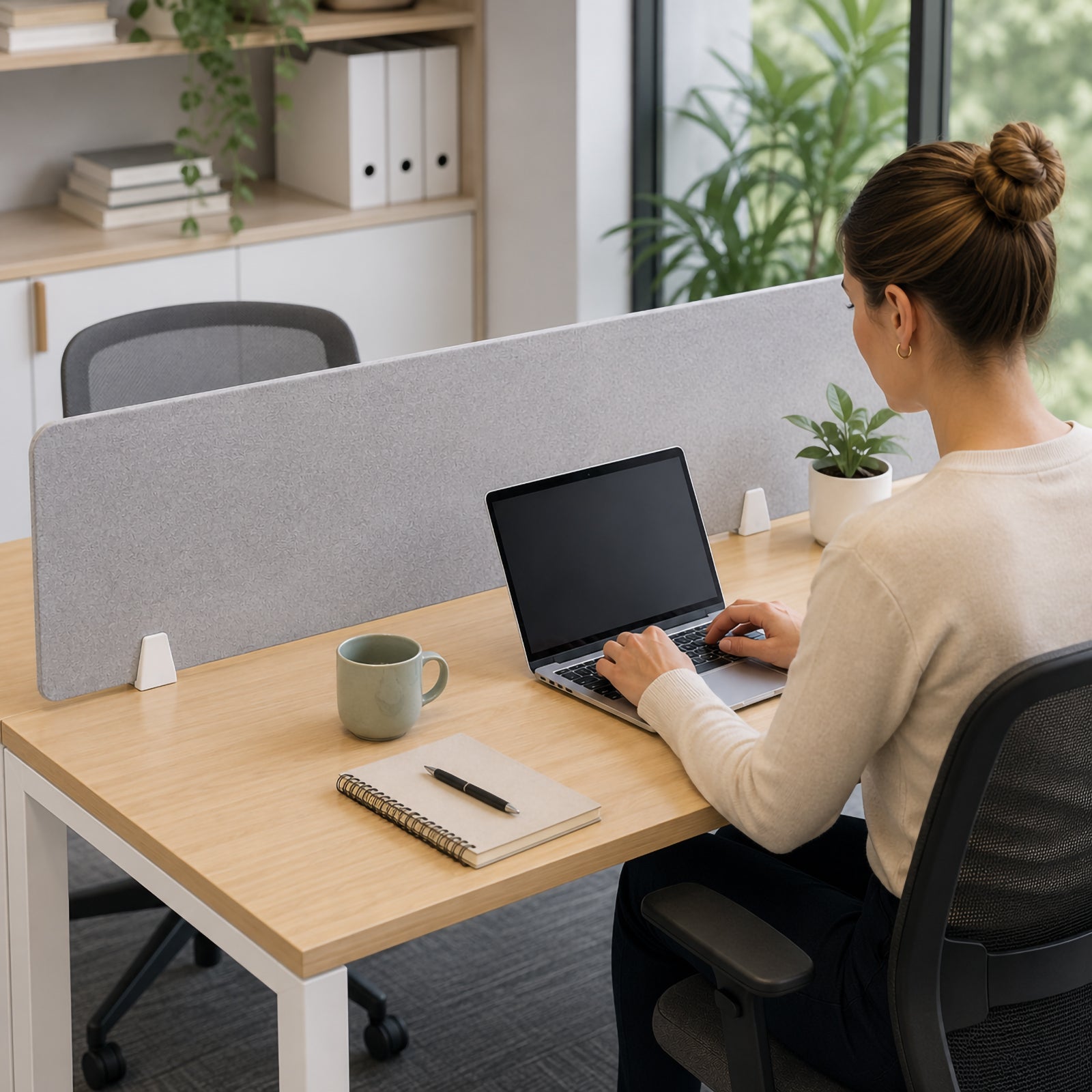 Visionchart SANA matte white desk screen clamps securing a grey PET privacy screen to a light timber office workstation while a person works on a laptop, showing the discreet white hardware coordinating with the desk frame in a bright modern office.