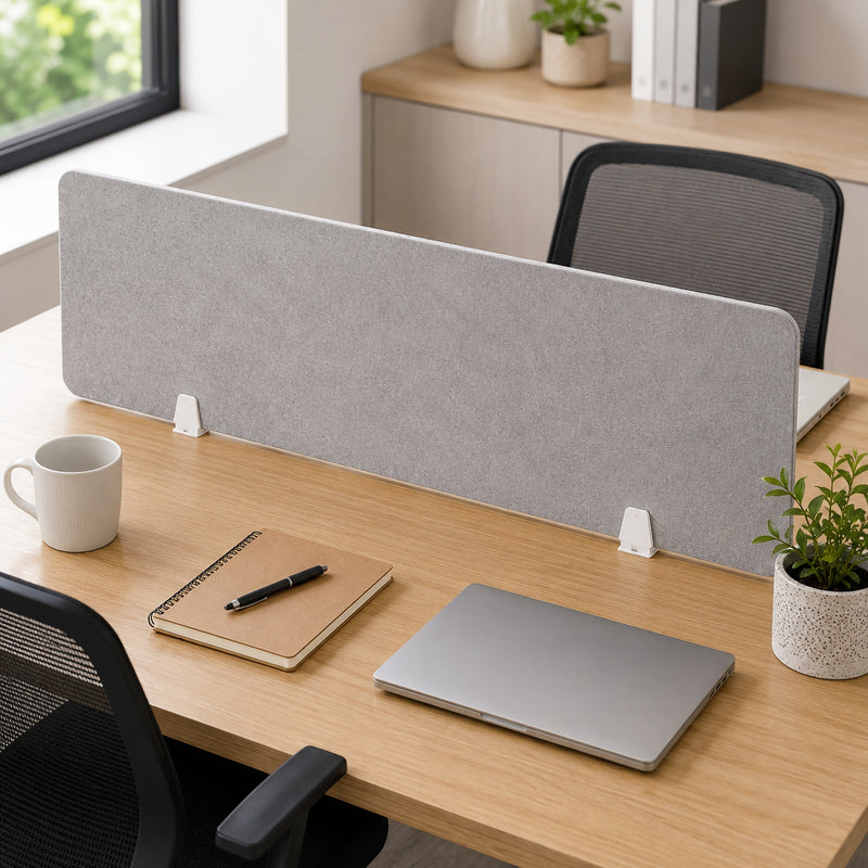 Grey PET privacy screen mounted across a contemporary timber office desk using two Visionchart SANA matte white desk screen clamps, with laptop, notebook, coffee mug and plants illustrating a neat shared workstation installation.