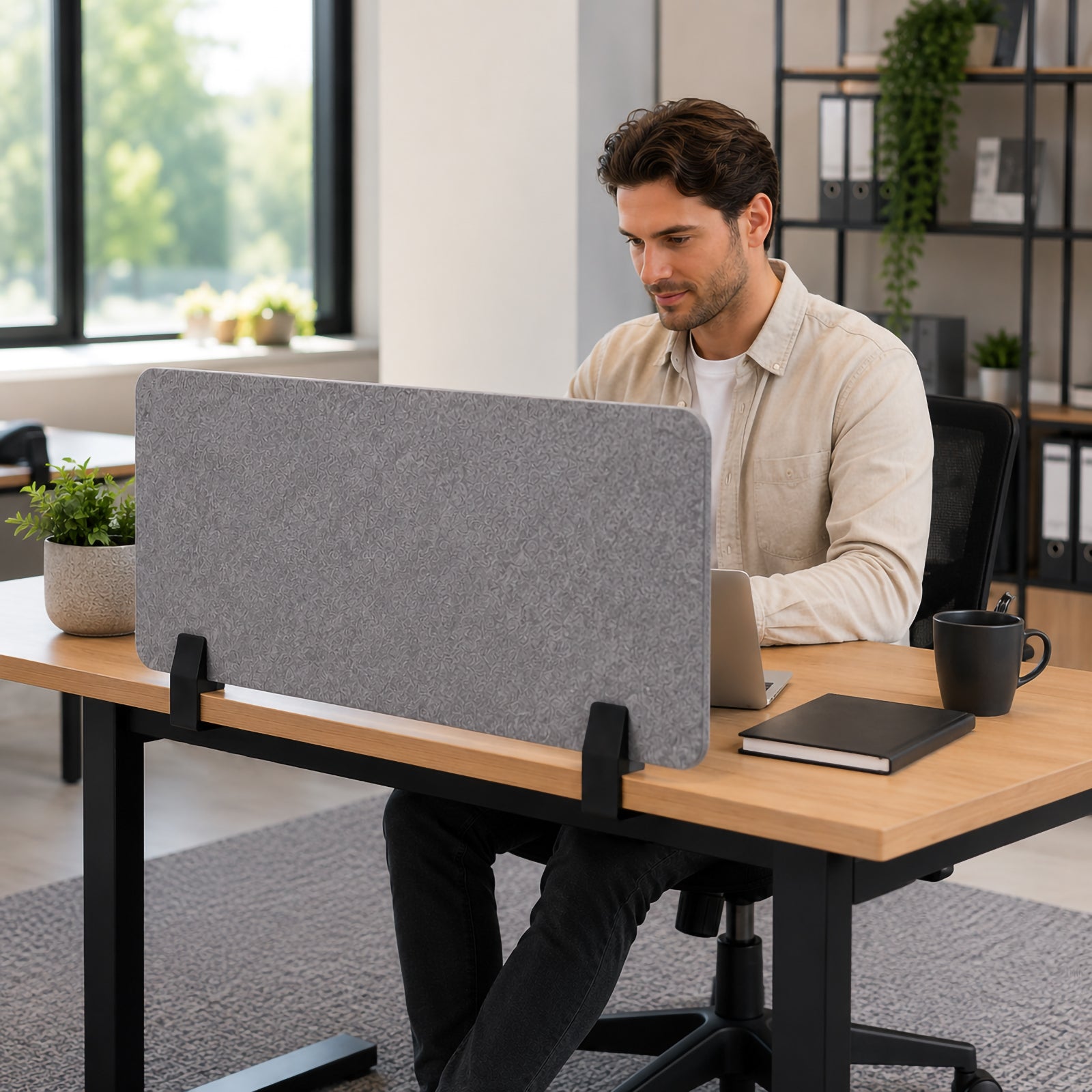 Visionchart SANA Type 2 matte black side mount clamps securing a grey PET acoustic desk screen to a timber workstation while a person works on a laptop in a bright contemporary office.
