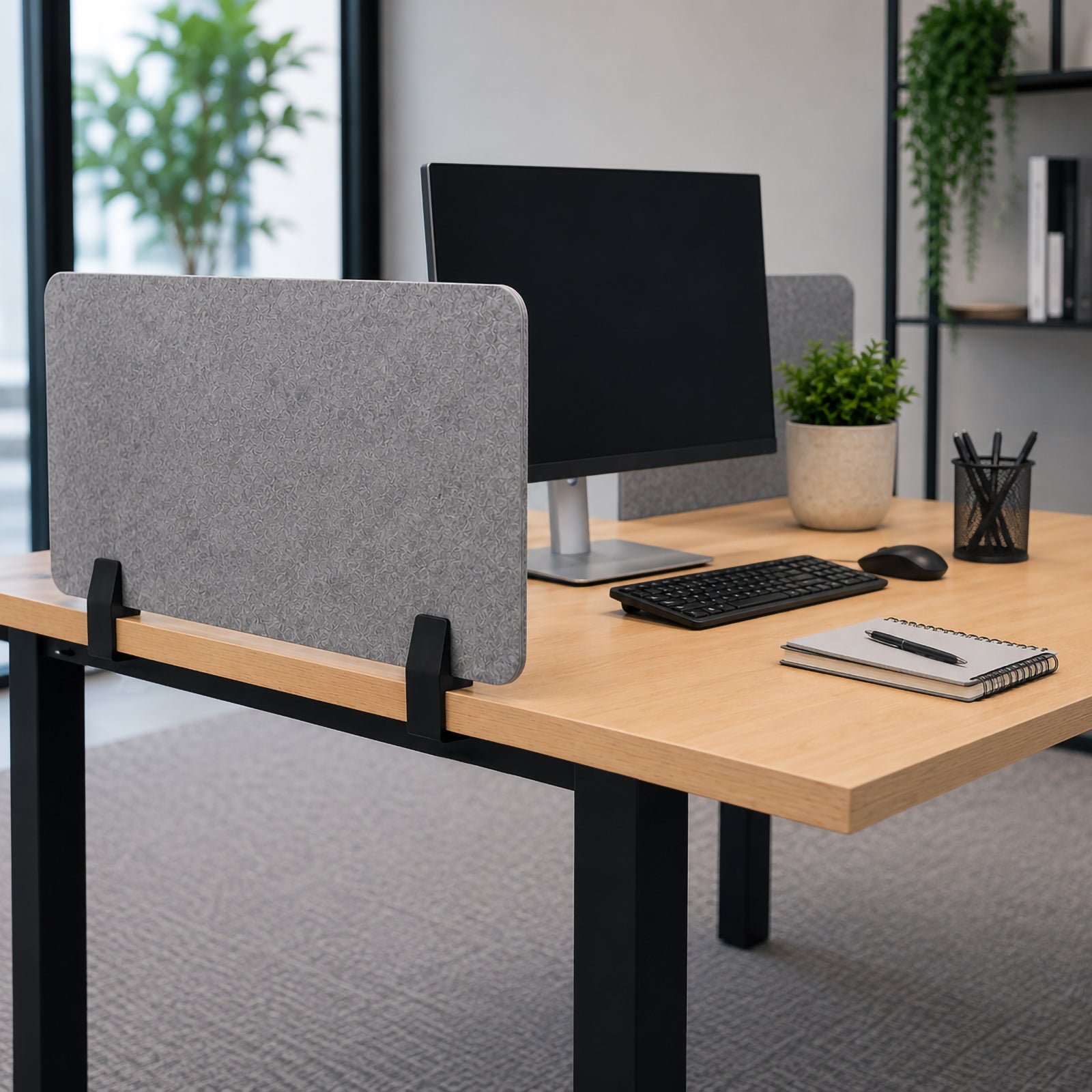 Modern workstation with grey PET acoustic desk screen secured by matte black SANA Type 2 side mount clamps, with monitor, keyboard, mouse, notebook and office plants arranged on a timber desk.