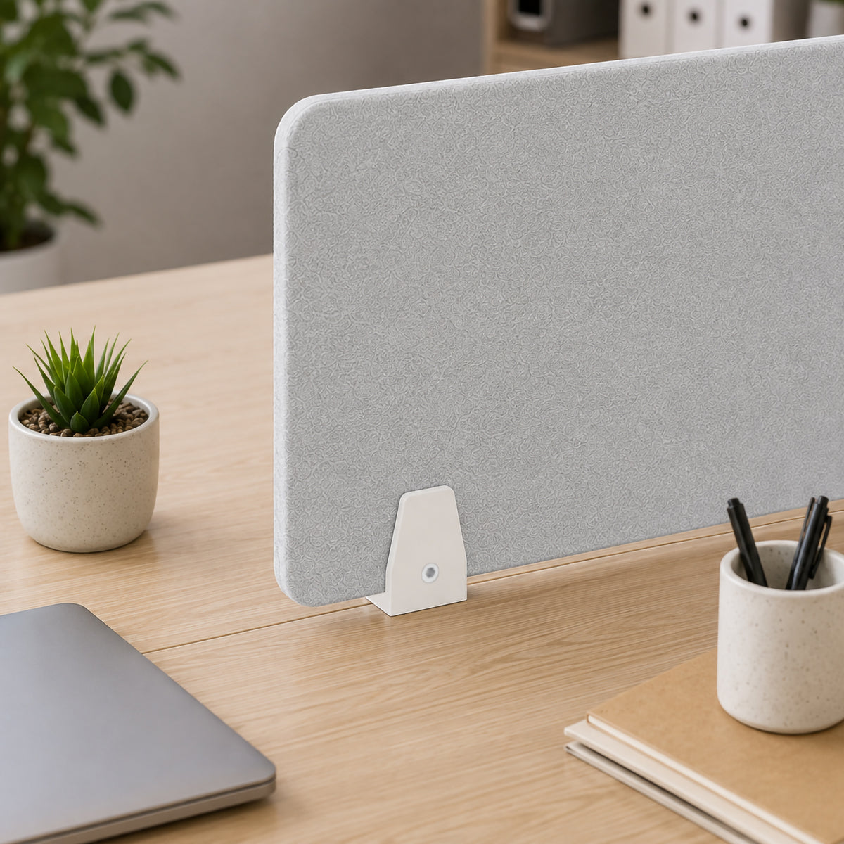 Close up of a SANA Type 3 matte white divide mount clamp holding a textured light grey PET acoustic screen between adjoining timber desktops, surrounded by a laptop, small plant, stationery holder and notebooks in a modern office setting.