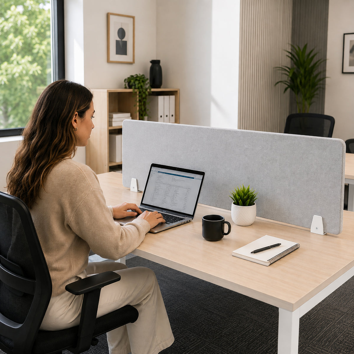 Visionchart SANA Type 3 matte white divide mount clamps securing a light grey PET acoustic screen between shared timber office desks while a woman works on a laptop, showing a bright contemporary workstation designed for privacy and focused work.
