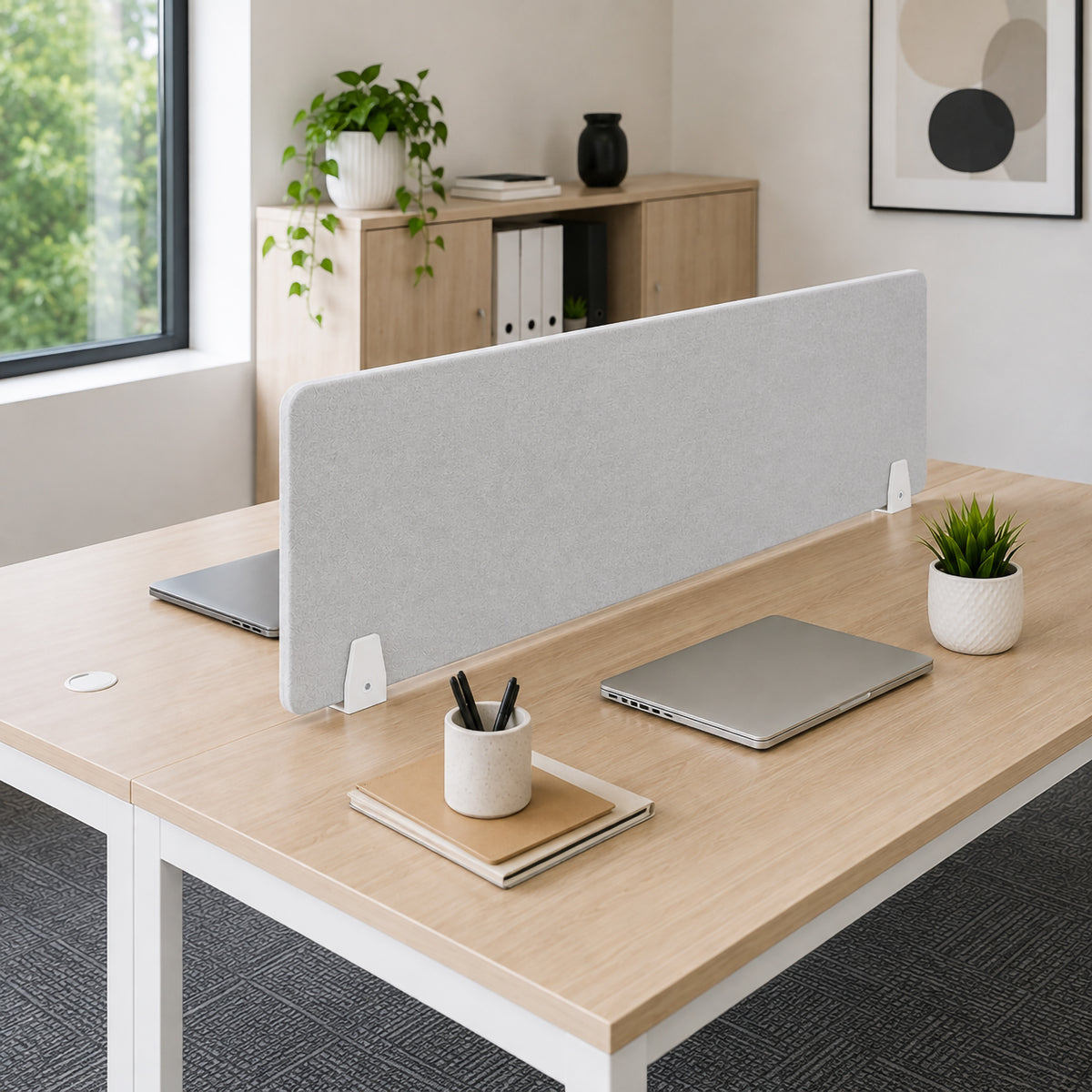 Wide lifestyle view of a light grey PET acoustic divider installed between two back to back timber workstations using Visionchart SANA Type 3 matte white clamps, with laptops, stationery and plants arranged across a bright shared office environment.