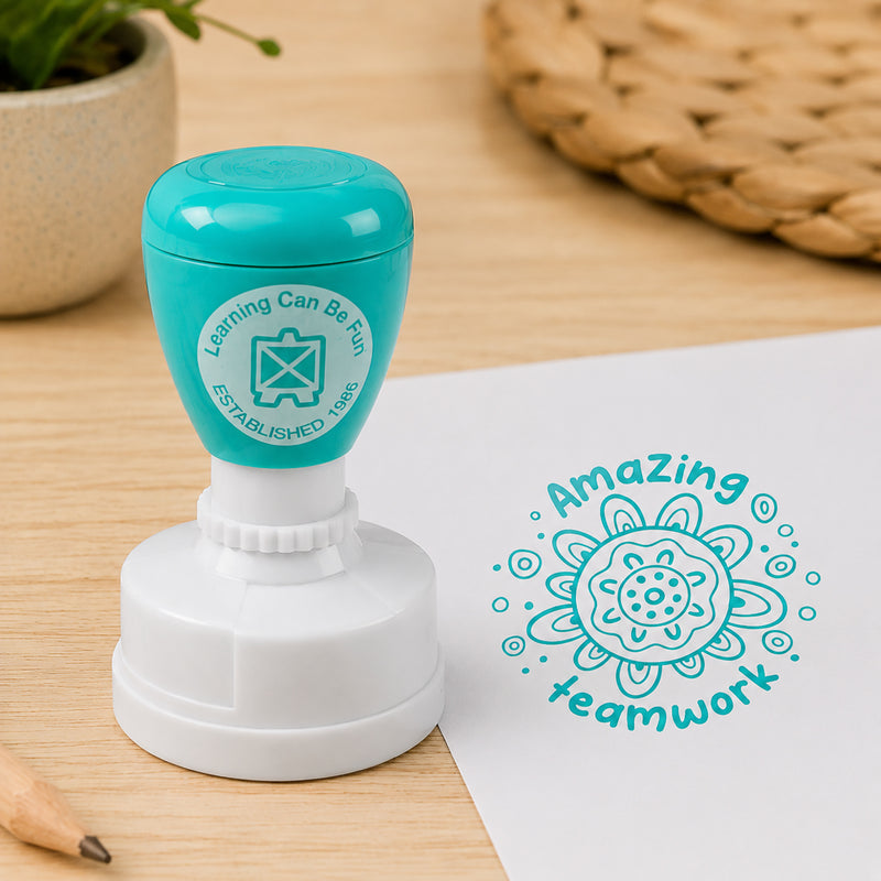 The self inking merit stamp sits beside a worksheet displaying a crisp Amazing Teamwork impression. Natural classroom elements including pencils and greenery highlight the stamp's professional quality and suitability for everyday educational use.