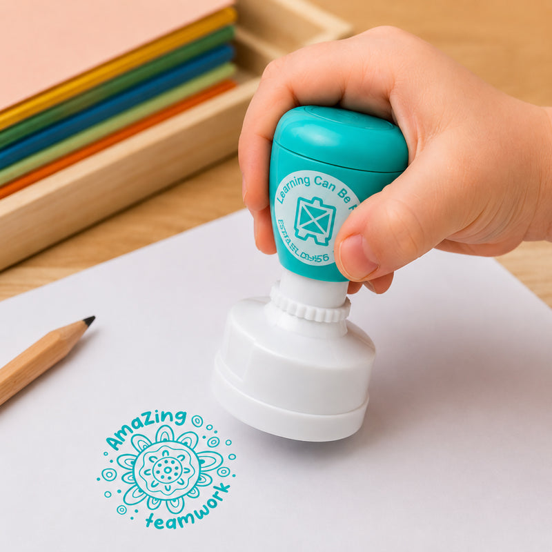 A child uses the Sacred Country Meeting Place Merit Stamp to create an Amazing Teamwork impression on a worksheet. The vibrant turquoise stamp showcases detailed Meeting Place artwork and demonstrates how teachers can provide positive feedback that celebrates collaboration and group achievement.