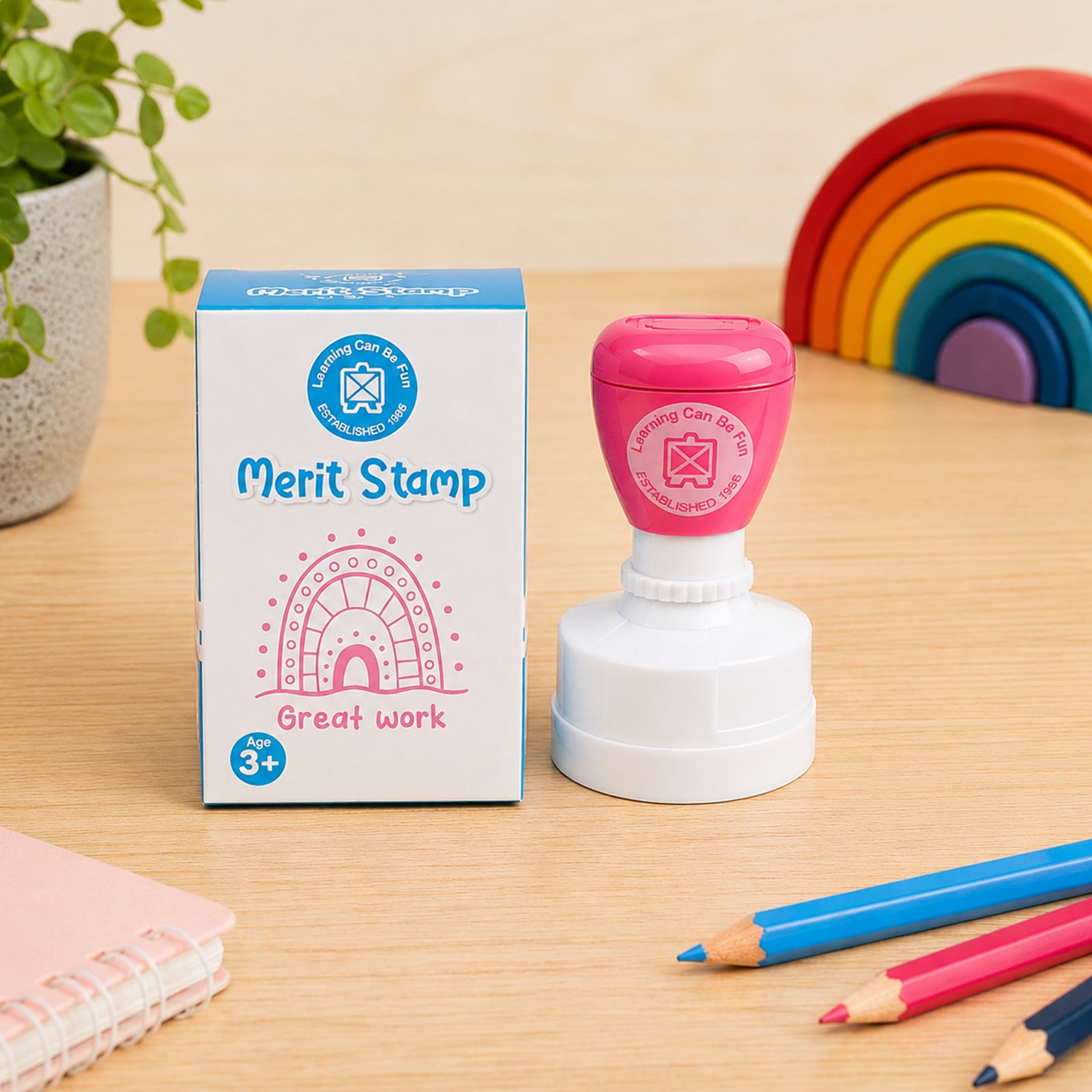 Product packaging and the Sacred Country Sandhill Merit Stamp are presented together on a classroom desk. The image highlights the Great Work message, colourful design and attractive retail presentation, making it ideal for teachers and educators.