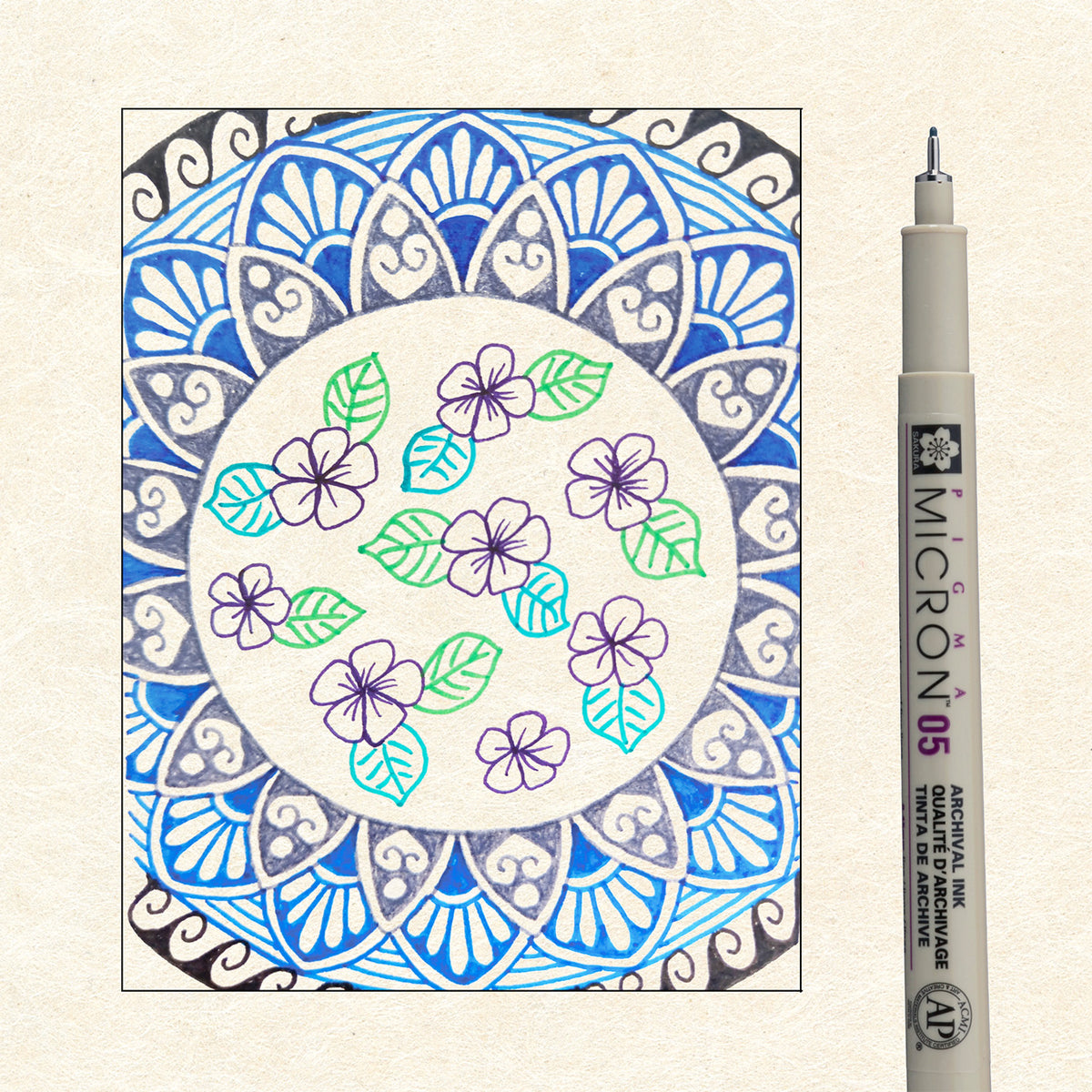 Sakura Pigma Mandala Cool Tone Set 05: blue mandala with purple flowers and green leaves drawn in Pigma ink beside a single Micron 05 pen