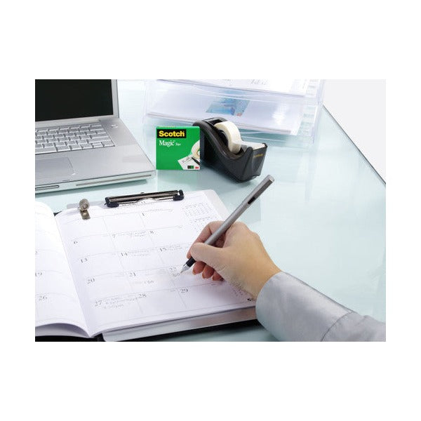 Person writing in a planner at a white desk beside a laptop, with a black Scotch tape dispenser and a green Scotch box