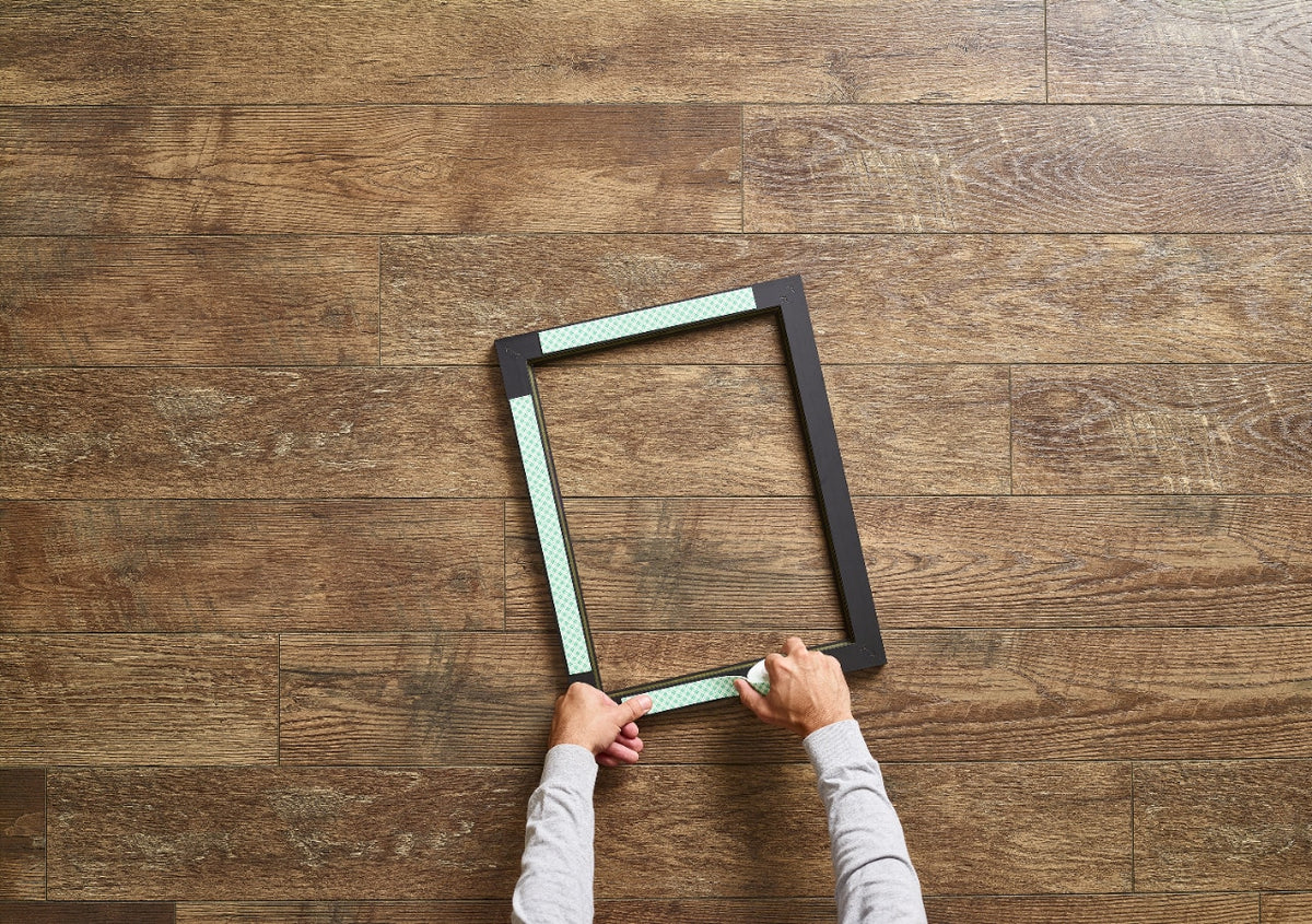 Hands pressing strips of Scotch-Mount indoor mounting tape onto the back of a black picture frame on a timber floor