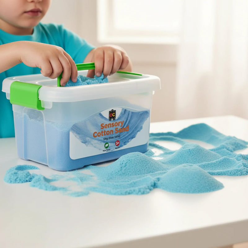 Child using Educational Colours Sensory Cotton Sand 5kg Blue, lifting soft blue sensory sand from a reusable storage tub while sand is spread across a white tabletop.