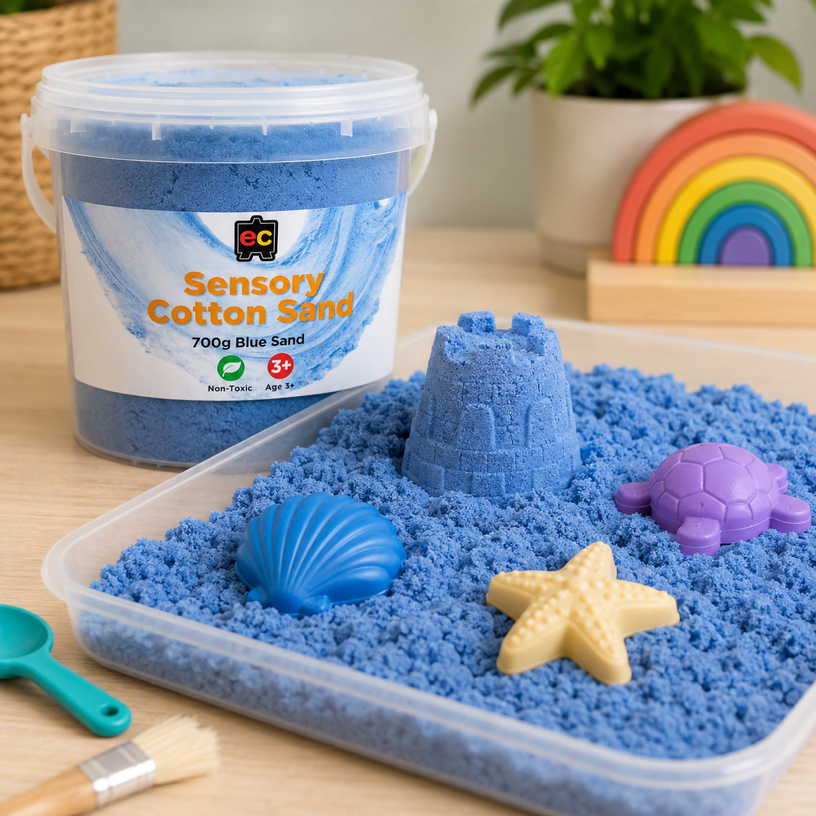 Blue sensory cotton sand displayed in a play tray with moulds, shells, and creative sensory play accessories beside a reusable container.