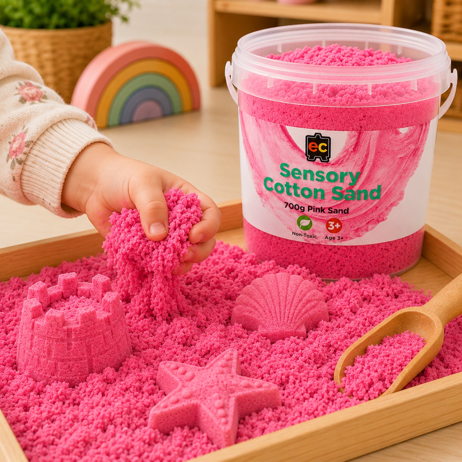Child stretching vibrant pink sensory cotton sand inside a wooden tray with moulded shell, star, and castle shapes beside Educational Colours storage tub.